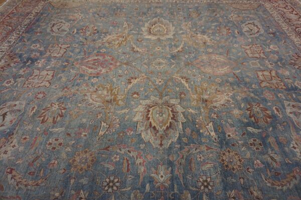 A distressed blue rug displays an intricate traditional floral and vine pattern in muted beige and rust tones.