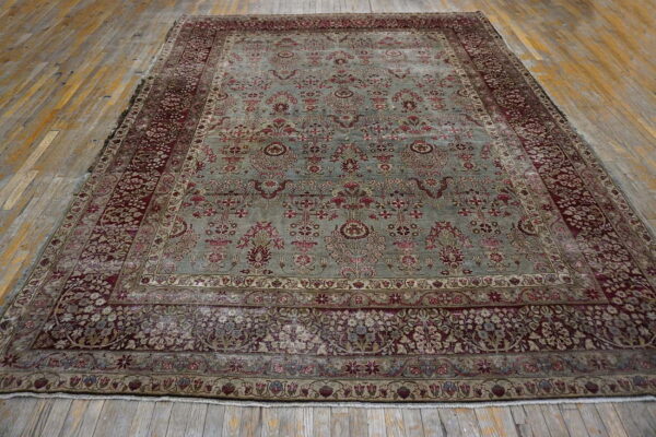 Ornate traditional rug with sage green field and burgundy floral borders displayed on wide wood planks.