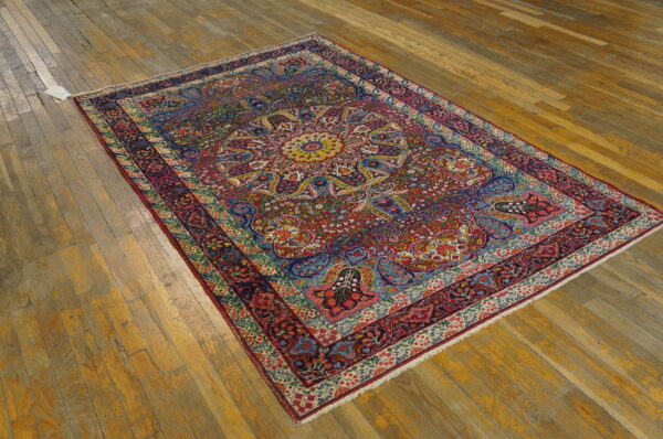 Jewel-toned rug with red, blue, and gold floral patterns lies on worn wide-plank wood flooring.