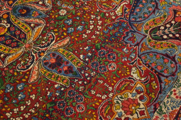 Detailed view of a low-pile rug with dense red, blue, yellow, and white floral and paisley motifs.