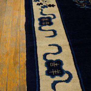 A close-up shows the navy rug border, featuring beige and blue winding motifs, next to distressed hardwood planks.