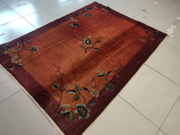 A reddish-orange rug with a low pile floral design and maroon border lies on white square tiles.