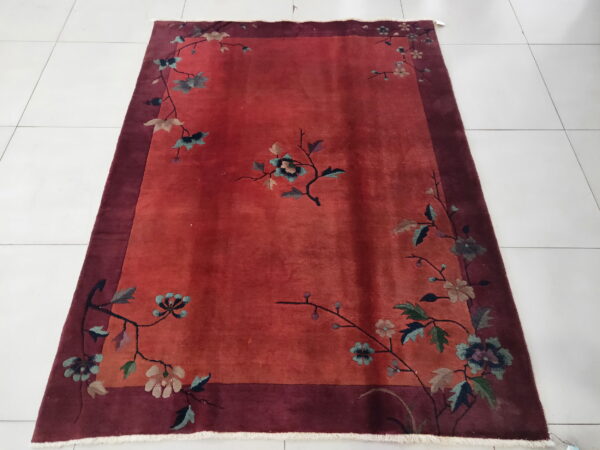 Low pile red rug with flowering branches and a wide maroon border on a bright white tiled floor.
