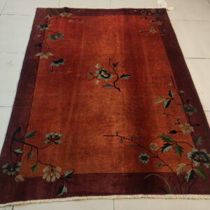 A reddish-orange rug with a deep maroon border, featuring muted floral vines, is displayed on white tile.