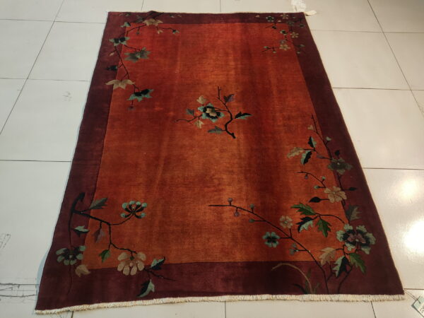 A reddish-orange rug with a deep maroon border, featuring muted floral vines, is displayed on white tile.