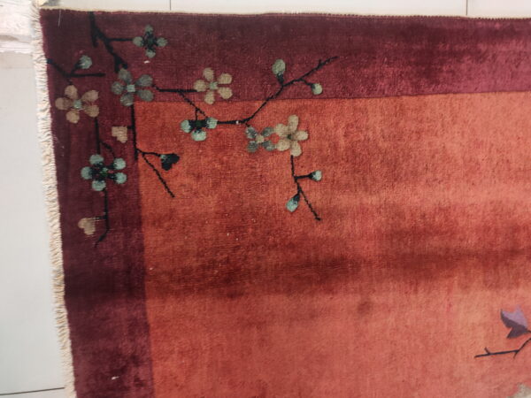 Shaded reddish-orange rug field with a maroon border and stylized floral pattern in muted blue and tan.