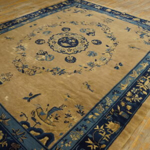 Tan field rug with detailed dark blue and teal floral and pictorial borders laid on wood floorboards.