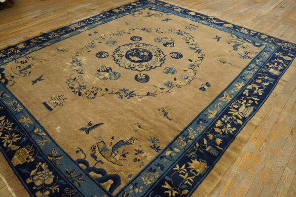 Tan field rug with detailed dark blue and teal floral and pictorial borders laid on wood floorboards.