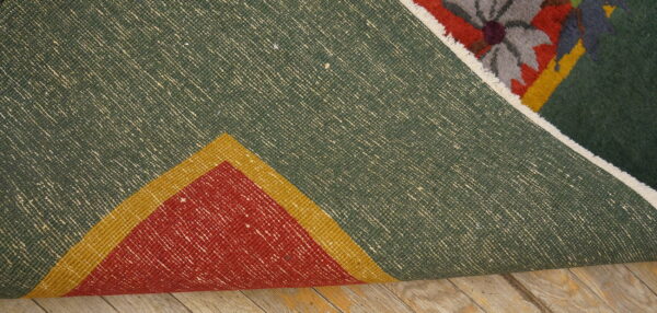 A rug is folded, showing its green, woven back and multicolored floral pile on distressed wooden flooring.