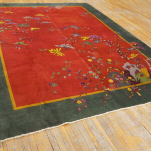 A low-pile rug with a red field, colorful floral vines, and a dark green border rests on distressed wood flooring.