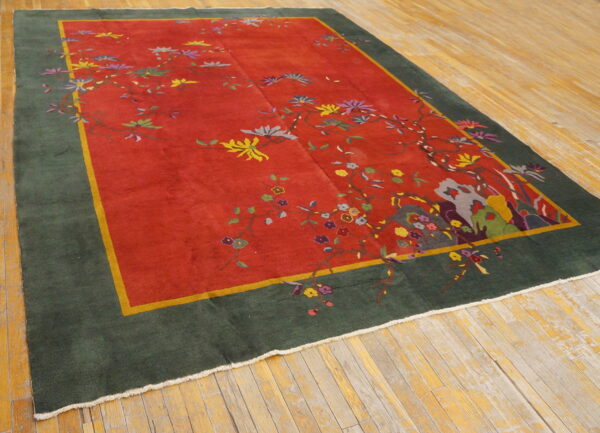 A low-pile rug with a red field, colorful floral vines, and a dark green border rests on distressed wood flooring.