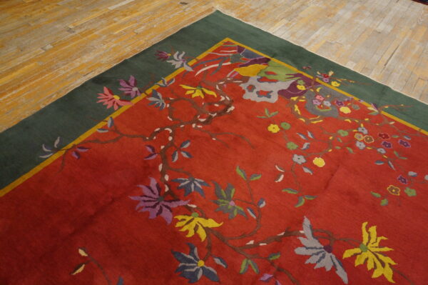 A red rug with a winding multi-colored floral vine pattern and dark green border rests on wood floors.