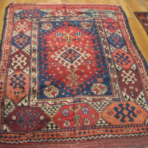 A low-pile rug displays intricate red, blue, and cream geometric patterns on light brown hardwood flooring.