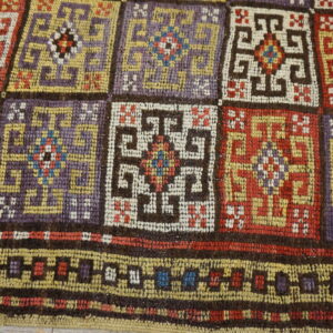 Coarse woven rug with geometric squares and a border in plum, red, gold, and cream colors.