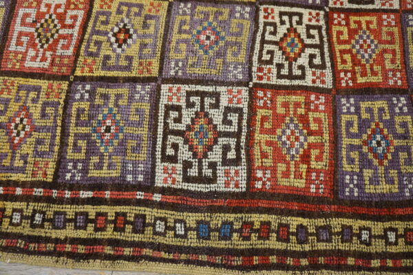 Coarse woven rug with geometric squares and a border in plum, red, gold, and cream colors.
