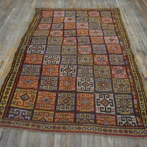Geometric checkerboard rug in red, purple, gold, and brown colors lying on gray distressed wood flooring.