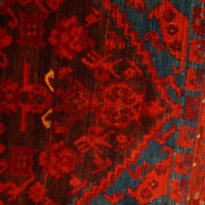 Close-up of a high-pile rug showing dense traditional motifs in bright red, dark brown, and teal blue.