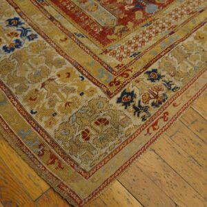 A corner of a traditional gold, red, and blue low-pile rug lies on distressed wood plank flooring.