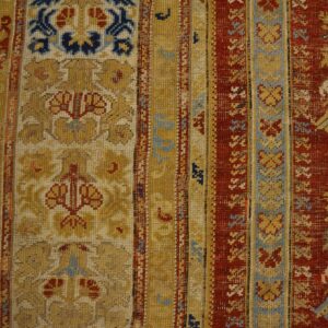 Close-up view of a rug border in red, gold, and blue complex patterns lying on wooden flooring.
