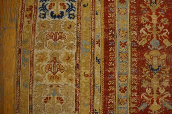 Close-up view of a rug border in red, gold, and blue complex patterns lying on wooden flooring.