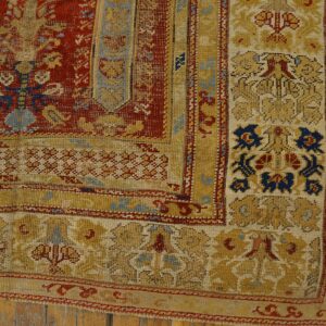 Distressed rug corner featuring dense red and gold patterns with blue details on wood flooring.