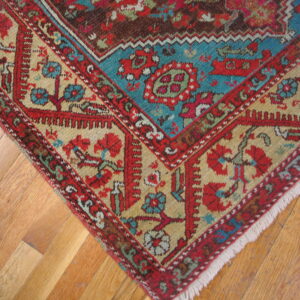 Red, teal, and gold patterned rug corner with fringes sits on light brown plank hardwood flooring.