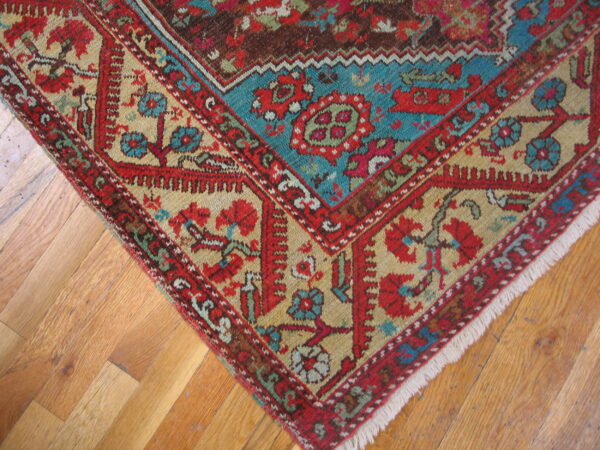 Red, teal, and gold patterned rug corner with fringes sits on light brown plank hardwood flooring.