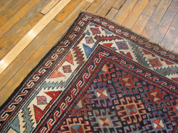 Corner detail of a woven rug with dense red, blue, and cream geometric patterns rests on diagonal hardwood flooring.