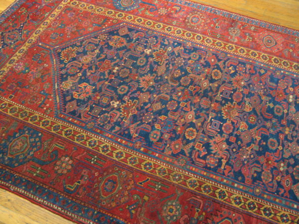 Close-up of a low-pile rug featuring a navy geometric field and detailed red borders on hardwood flooring.