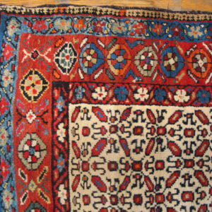 Red, blue, and cream geometric patterned rug showing corner details and fringes on wood flooring.