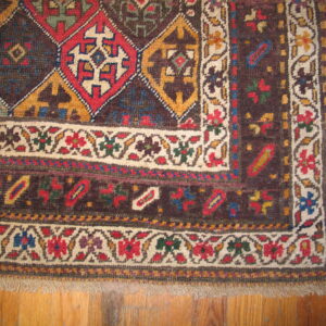 Low-pile rug corner showing dark brown geometric field and detailed floral borders on light brown hardwood.