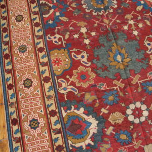 Close-up of a red rug with a complex multicolored border and floral field on natural hardwood flooring.