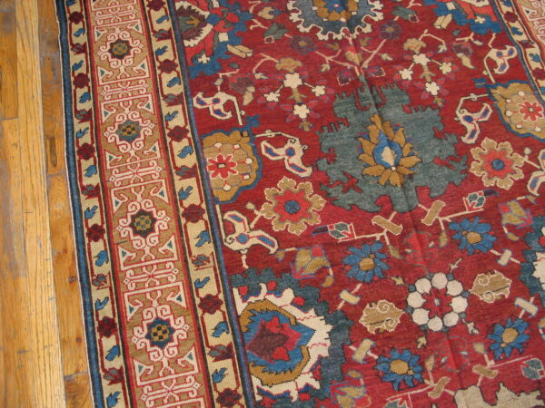Close-up of a red rug with a complex multicolored border and floral field on natural hardwood flooring.