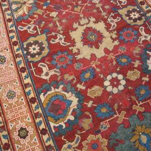 Ornate traditional rug in red, blue, and cream colors, showing wide border detail on wood flooring.