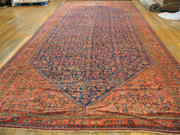 Oversized rug with a dense blue field and rust-red patterned spandrels rests on wood flooring.