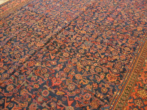 Navy rug with dense, repeating geometric motifs in orange, red, and teal, showing creases across the surface.