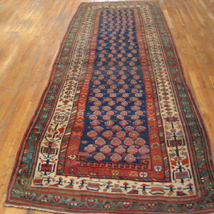 Narrow runner rug with a navy field, repeating red designs, and complex borders, lying on light wood flooring.