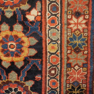Detail view of a rug showing a dark field and borders patterned with rust, blue, gold, and sage green motifs.