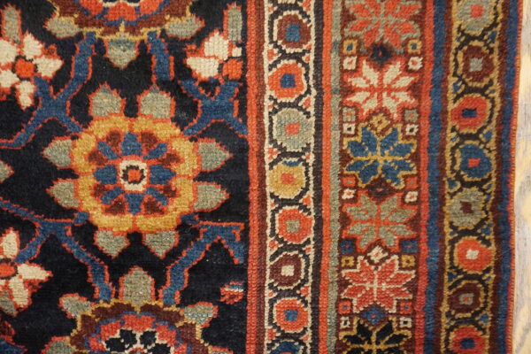 Detail view of a rug showing a dark field and borders patterned with rust, blue, gold, and sage green motifs.