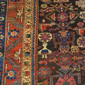 Geometric rug detail showing a dark brown field, red border, and a stylized figure on light wooden flooring.