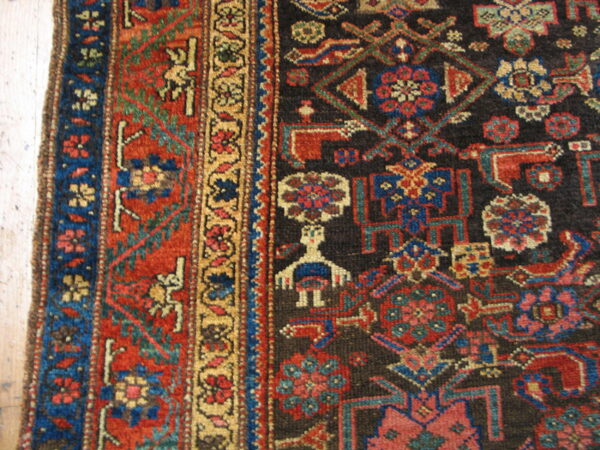 Geometric rug detail showing a dark brown field, red border, and a stylized figure on light wooden flooring.