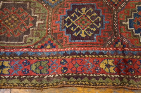 Geometric rug border and field in muted red, blue, green, and brown, laid on wood floorboards.