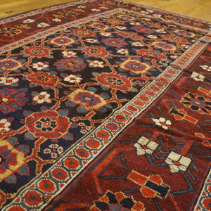 Deep red and navy rug with repeating stylized floral patterns and multiple geometric borders on hardwood flooring.