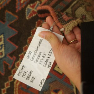 A hand holds a tag near the fringed edge of a geometric rug with brown, red, blue, and cream patterns.