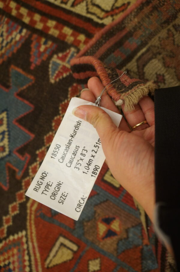 A hand holds a tag near the fringed edge of a geometric rug with brown, red, blue, and cream patterns.