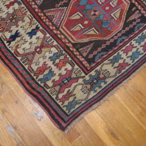 Intricate red, dark brown, blue, and beige patterned rug corner on light wood flooring.