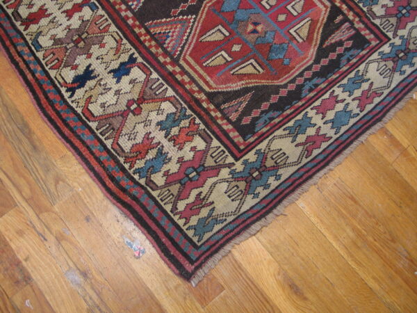Intricate red, dark brown, blue, and beige patterned rug corner on light wood flooring.