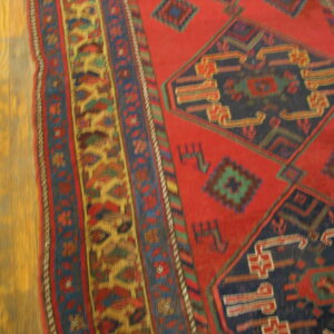 A red and navy geometric rug with detailed borders rests on golden-toned wood planks.