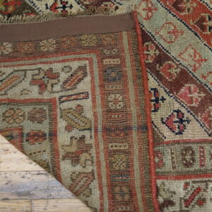 A folded corner of a geometric patterned rug in muted reds and greens rests on distressed wood flooring.