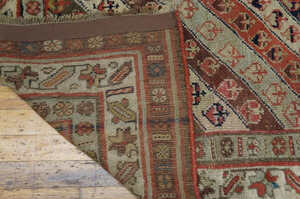 A folded corner of a geometric patterned rug in muted reds and greens rests on distressed wood flooring.